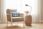 A white armchair with woven cane panels and a beige upholstered seat, styled with a blue and white patterned accent pillow. It is set in a bright room with white sheer curtains, a wooden floor, a matching woven side table, a gold floor lamp, and a vase of pink flowers.
