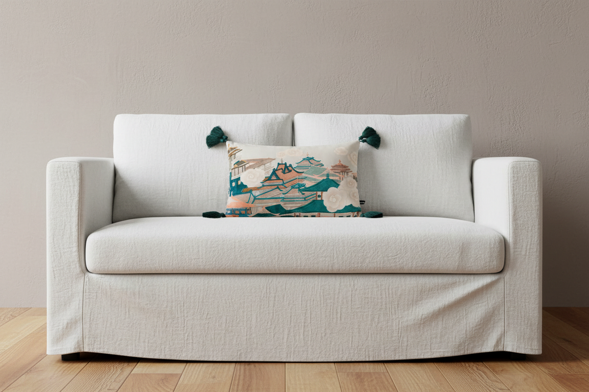 A beige upholstered armchair with woven cane side panels and white legs, styled in a bright room with a decorative multicolor landscape throw pillow, a patterned cream pillow, a gold floor lamp, and a wooden side table with pink flowers.