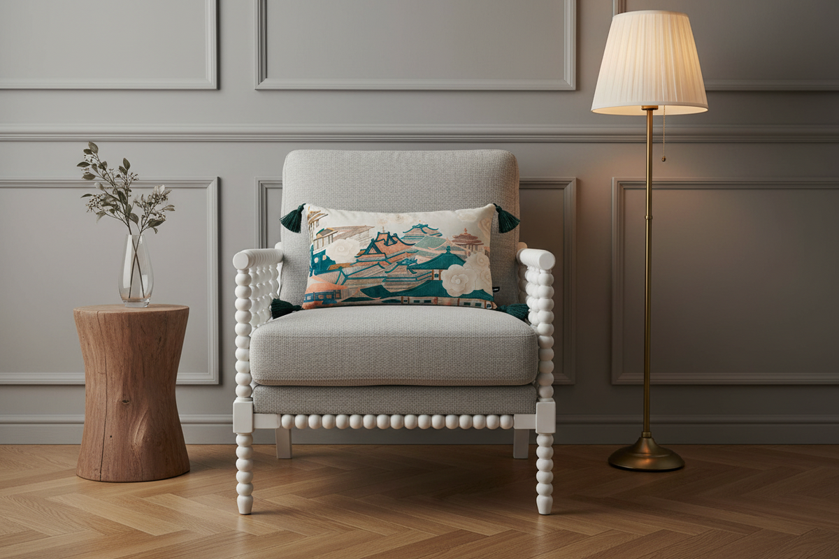 A beige upholstered armchair with woven cane side panels and white carved legs, styled in a bright room with a decorative multicolor landscape throw pillow, a patterned cream pillow, a gold floor lamp, and a wooden side table with pink flowers.