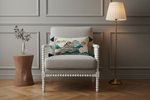 A beige upholstered armchair with woven cane side panels and white carved legs, styled in a bright room with a decorative multicolor landscape throw pillow, a patterned cream pillow, a gold floor lamp, and a wooden side table with pink flowers.