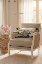 A beige upholstered armchair with woven cane side panels and white legs, styled in a bright room with a decorative multicolor landscape throw pillow, a patterned cream pillow, a gold floor lamp, and a wooden side table with pink flowers.