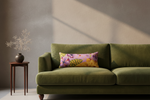 An olive green velvet sofa in a styled room setting, featuring a decorative rectangular throw pillow with a colorful butterfly and floral pattern on a pink background. A small wooden side table with a dark vase and dried branches stands to the left against a textured beige wall with soft window light shadows.