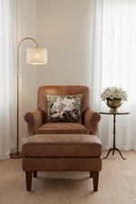 A brown leather armchair with a matching ottoman in a bright, neutral room. A floral throw pillow rests on the chair, and a gold floor lamp, a small side table with a vase of white flowers, and sheer white curtains frame the scene.