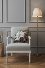 A light gray upholstered armchair with white turned legs sits in a styled room with gray paneled walls, a floor lamp, wood flooring, and a floral accent pillow.
