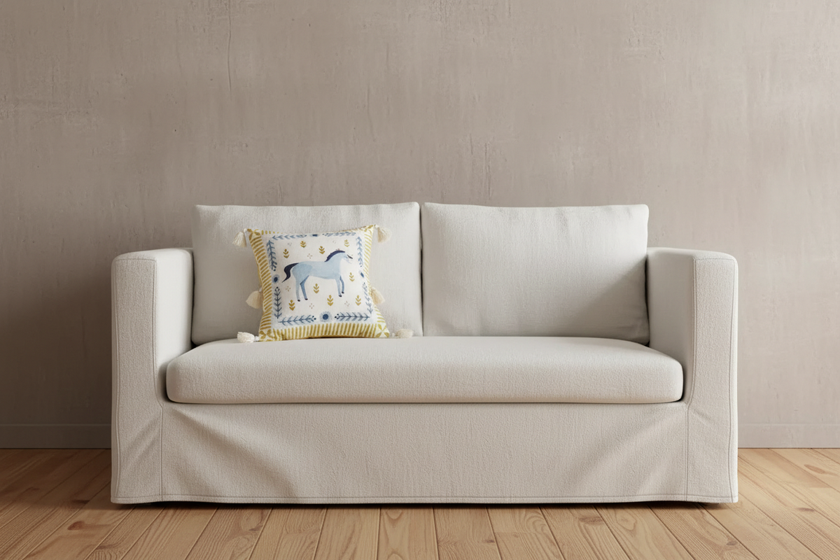 A cream-colored slipcovered loveseat sofa with a decorative throw pillow featuring a blue horse design, set against a textured beige wall and light wood flooring.