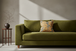 An olive green velvet sofa in a modern room setting with a decorative yellow and blue patterned throw pillow, a small wooden side table with a dark vase of branches, and dramatic diagonal sunlight casting shadows on the wall.