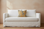 An olive green velvet sofa in a modern room setting with a decorative yellow and blue patterned throw pillow, a small wooden side table with a dark vase of branches, and dramatic diagonal sunlight casting shadows on the wall.