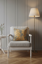 An olive green velvet sofa in a modern room setting with a decorative yellow and blue patterned throw pillow, a small wooden side table with a dark vase of branches, and dramatic diagonal sunlight casting shadows on the wall.