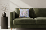 An ornate square throw pillow with a detailed blue, gold, and pink damask-style pattern, resting on a green velvet sofa next to a dark side table with a vase of branches.