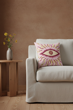A square throw pillow with a stylized pink and yellow eye design, resting on a light beige sofa in a cozy room setting with a wooden side table and vase of flowers.