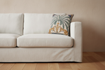 A beige upholstered rattan armchair in a bright room, styled with a square decorative throw pillow featuring a palm tree and two lions. A carved wooden side table with pink flowers and a gold floor lamp sit beside the chair.