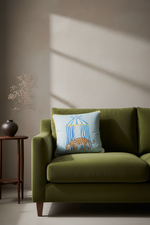 A cream-colored slipcovered sofa in a styled living room setting, featuring a light blue decorative throw pillow with a birdcage illustration, next to a wooden coffee table and a potted plant.
