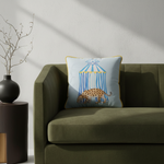 A cream-colored slipcovered sofa in a styled living room setting, featuring a light blue decorative throw pillow with a birdcage illustration, next to a wooden coffee table and a potted plant.