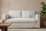 A cream-colored slipcovered sofa in a styled living room setting, featuring a light blue decorative throw pillow with a birdcage illustration, next to a wooden coffee table and a potted plant.
