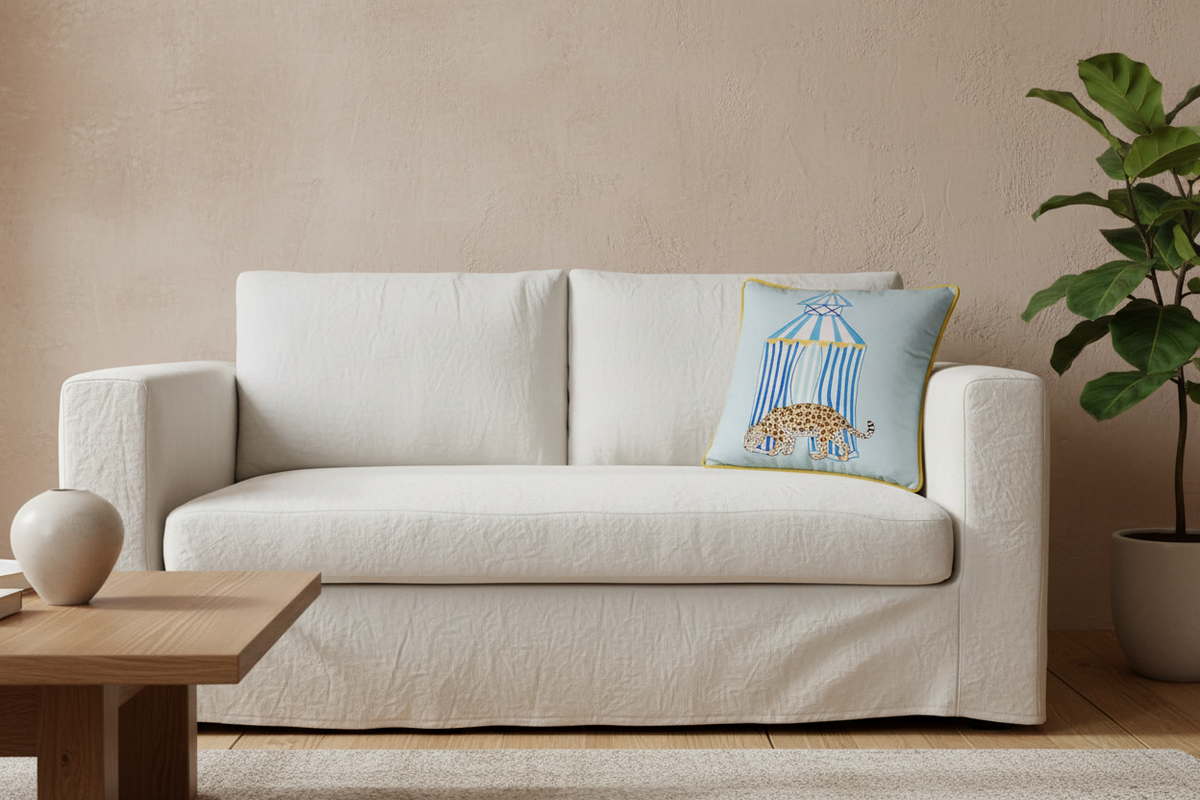 A cream-colored slipcovered sofa in a styled living room setting, featuring a light blue decorative throw pillow with a birdcage illustration, next to a wooden coffee table and a potted plant.