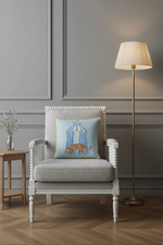 A cream-colored slipcovered sofa in a styled living room setting, featuring a light blue decorative throw pillow with a birdcage illustration, next to a wooden coffee table and a potted plant.