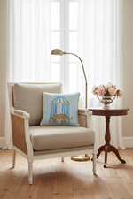 A cream-colored slipcovered sofa in a styled living room setting, featuring a light blue decorative throw pillow with a birdcage illustration, next to a wooden coffee table and a potted plant.