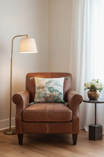 A white fabric slipcovered sofa in a minimalist room, styled with a square decorative throw pillow featuring a traditional East Asian landscape print and green pom-poms.