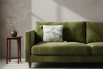 An olive green velvet sofa in a modern living room setting, styled with a square throw pillow featuring a traditional Chinese landscape print. A small wooden side table with a vase of dried branches sits to the left of the sofa against a textured gray wall.