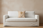 A cream-colored slipcovered loveseat sofa with a decorative beige throw pillow featuring a gold sun and green botanical pattern, set against a textured beige wall and light wood floor.