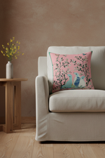 A square decorative throw pillow with a pink background and a bird-and-branch print, styled on a cream-colored armchair in a warm, minimalist room setting.