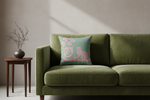 A square decorative throw pillow with a pink cat and floral design on a mint green background, styled on an olive green velvet sofa next to a small wooden side table.