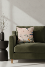 A square throw pillow with a light pink background and a traditional Chinese-style bird and landscape print, resting on an olive green velvet sofa in a modern living room setting.