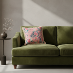 An olive green velvet loveseat sofa in a styled room setting, featuring a decorative pink floral throw pillow with a fringed edge.