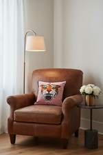 A brown leather armchair in a styled room setting, featuring a pink throw pillow with a tiger face design. A gold floor lamp stands to the left, and a small side table with white roses is on the right.