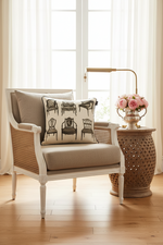 A white and cane armchair with beige cushions sits in a bright room beside a patterned accent pillow, a decorative side table with a lamp and pink flowers, and sheer curtains over a window.