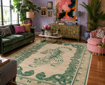 A large rectangular area rug with a cream background and a green tiger and scroll border, shown in a styled living room with a green sofa, pink armchair, cabinet, and plants.