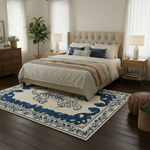 A styled bedroom scene showing a large rectangular area rug with a cream background and bold blue border and tiger motif, placed under a bed in a real interior with wood flooring, nightstands, lamps, and a plant.