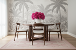 Dining room with a round wooden table and chairs, pink rug, flowers in a vase, and palm tree wallpaper.