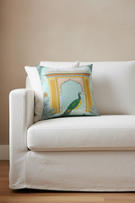 A decorative throw pillow featuring a turquoise and gold architectural arch design with a peacock, styled on a white slipcovered sofa against a beige wall.
