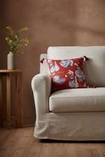 An off-white linen slipcovered loveseat sofa with a red patterned accent pillow, set against a textured beige wall and light wood flooring.