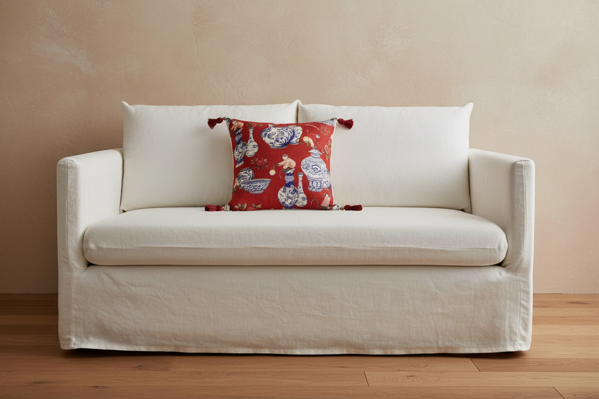 An off-white linen slipcovered loveseat sofa with a red patterned accent pillow, set against a textured beige wall and light wood flooring.