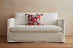 An off-white linen slipcovered loveseat sofa with a red patterned accent pillow, set against a textured beige wall and light wood flooring.