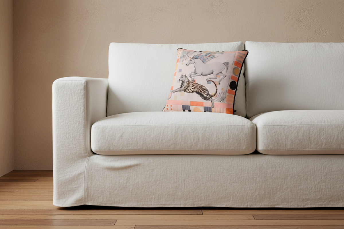 A white slipcovered sofa with a decorative throw pillow featuring a colorful animal print, set against a beige wall and light wood floor.