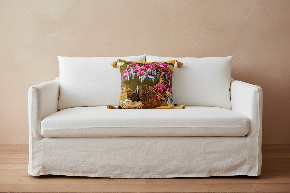 A white slipcovered loveseat sofa with a decorative square throw pillow featuring a colorful forest and bird design, set against a warm beige wall and light wood floor.