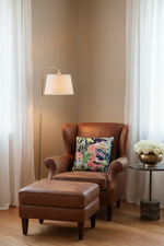 A light gray upholstered armchair with a colorful tropical floral pillow, set in a styled room with a wooden floor, a floor lamp, and a side table.