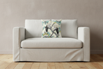 A light beige slipcovered loveseat sofa with a decorative throw pillow featuring a Japanese-style crane print, set against a textured beige wall and light wood flooring.