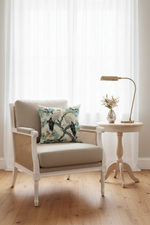 A light beige slipcovered loveseat sofa with a decorative throw pillow featuring a Japanese-style crane print, set against a textured beige wall and light wood flooring.