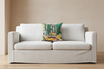 A cream-colored fabric slipcovered loveseat sofa with a colorful, illustrated throw pillow, set against a warm beige wall and light wood floor.