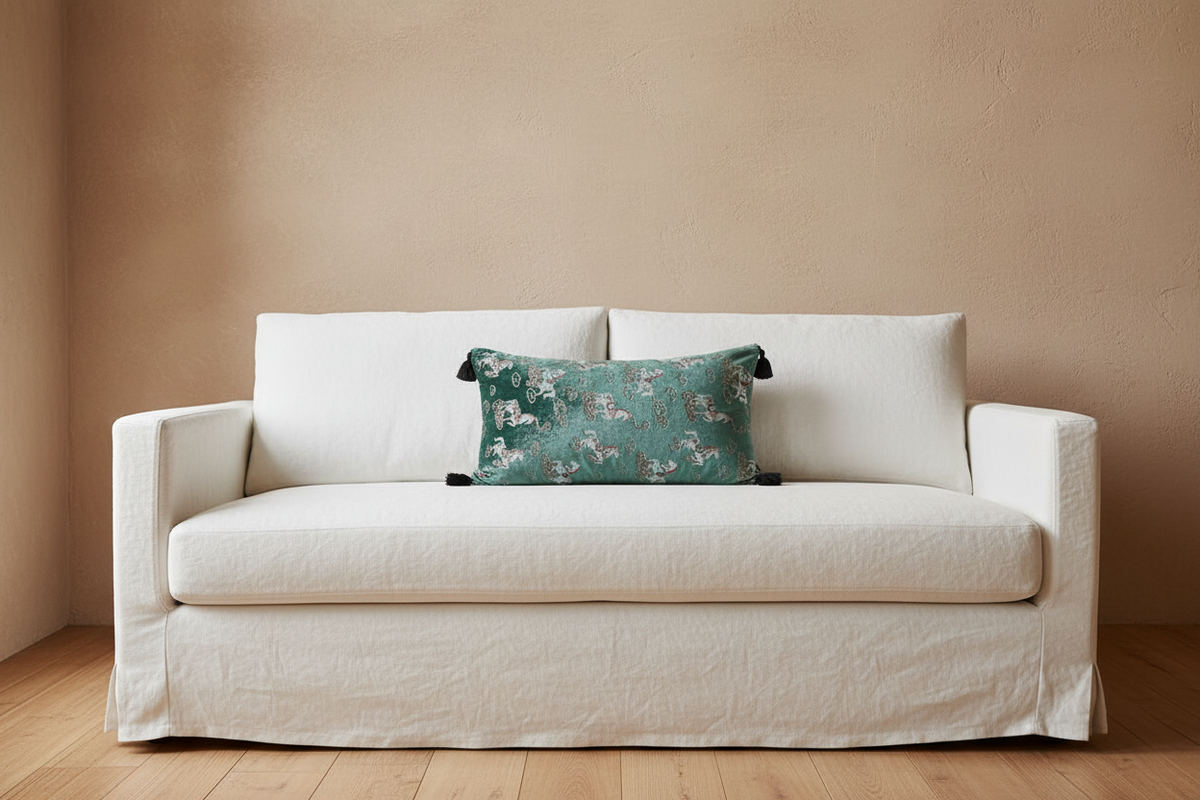 A cream-colored slipcovered loveseat sofa with a textured beige wall and light wood floor, styled with a green floral accent pillow.