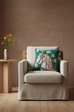 A beige upholstered armchair in a styled room setting, featuring a decorative square throw pillow with a green and white horse-and-carriage pattern. A small wooden side table with yellow flowers stands to the left against a textured brown wall and light wood floor.