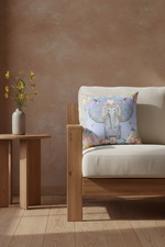 A cream-colored slipcovered loveseat sofa with two seat cushions and a decorative square throw pillow featuring a colorful elephant illustration, set against a beige wall and light wood floor.