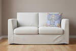 A cream-colored slipcovered loveseat sofa with two seat cushions and a decorative square throw pillow featuring a colorful elephant illustration, set against a beige wall and light wood floor.