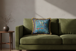 A square decorative throw pillow with a colorful landscape print and gold fringe, styled on an olive green velvet sofa next to a wooden side table.