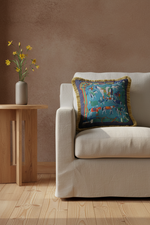A square decorative throw pillow with a colorful landscape print and gold fringe, styled on an olive green velvet sofa next to a wooden side table.