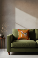 An olive green velvet sofa in a styled room setting, featuring a square orange throw pillow with a white embroidered horse and fringe trim.